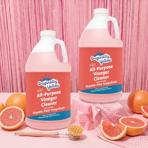 Two large jugs of Originally Yellow All-Purpose Vinegar Cleaner in Organic Pink Grapefruit flavor, displayed on a pink surface with grapefruit slices and whole grapefruits, set against a pink striped background.